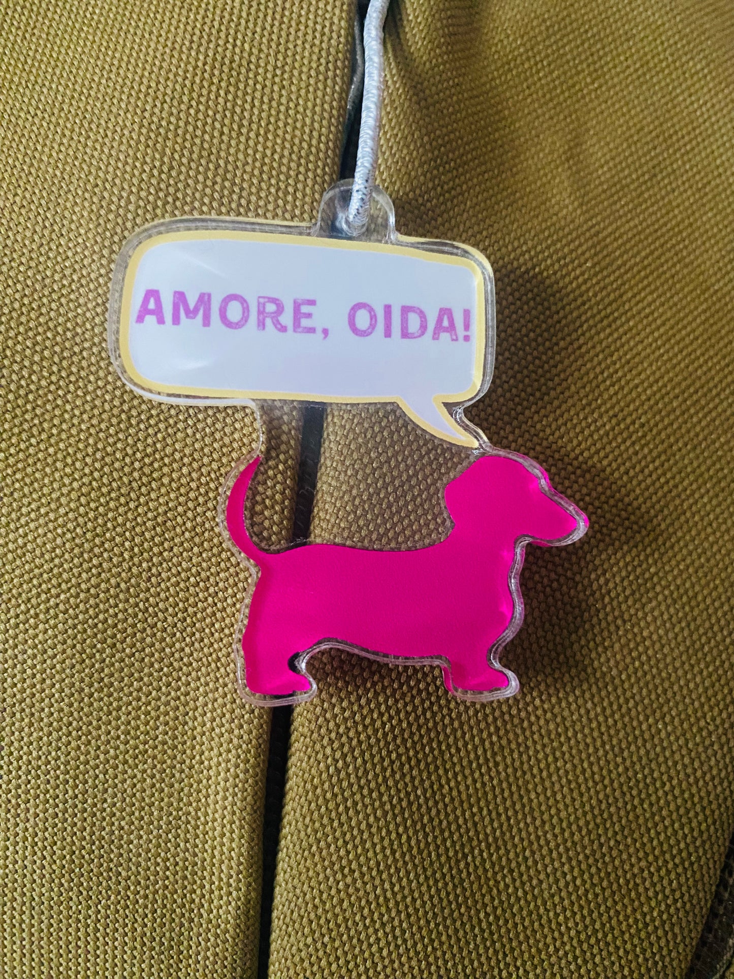 Anhänger aus Acryl in Form eines Dackels, Farbe pink. Drüber eine Sprechblase mit den Worten "Amore, Oida!". Der Anhänger ist mit einem Band versehen und der Hintergrund ist ein grünes Gewebe.