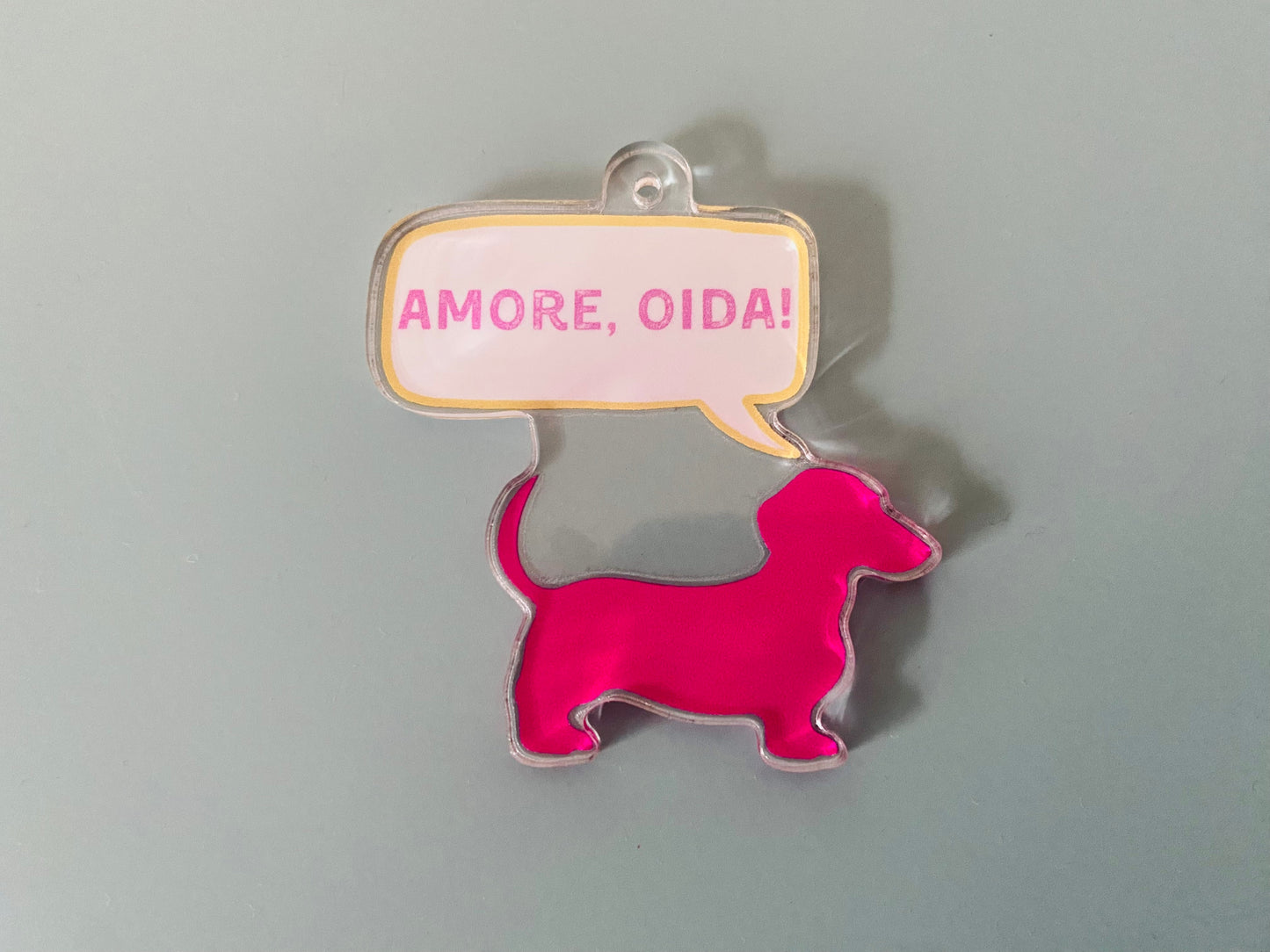 Acrylform in Form eines Dackels in pink, mit Sprechblase und den Worten Amore, Oida!. Oben mit Loch zum Aufhängen versehen.
