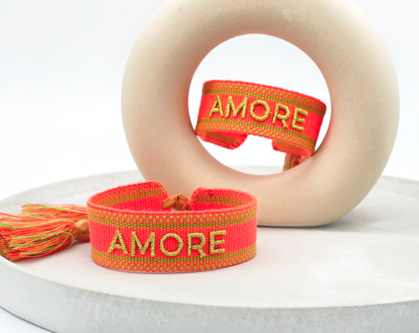 Gewebtes Armband in orange mit goldener Aufschrift "Amore" auf weißem Untergrund mit Quasten aif helle runden Stein liegend.