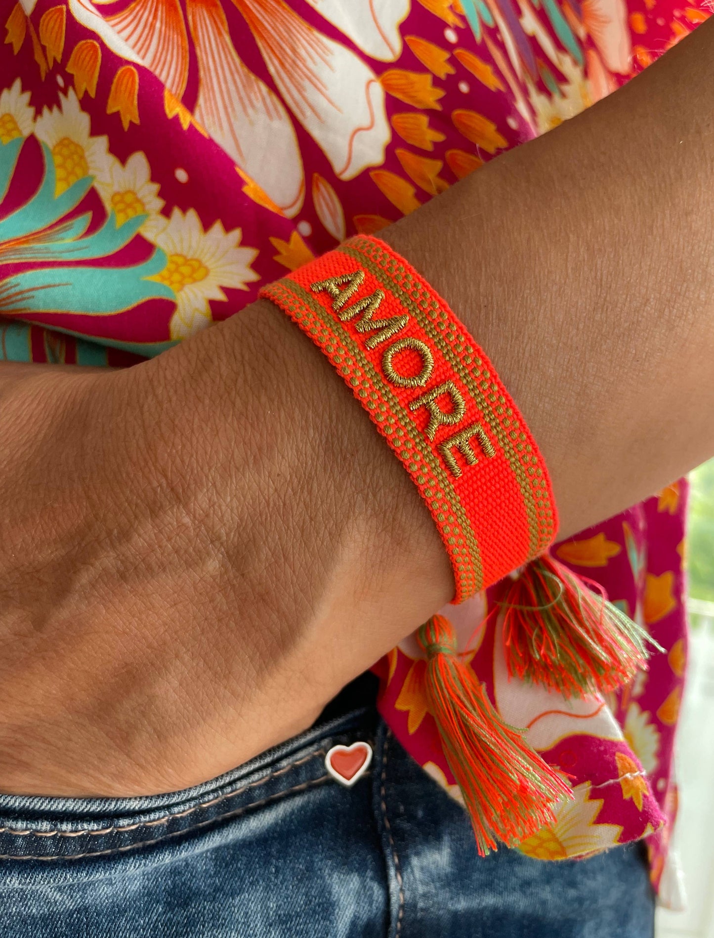 Gewebtes Armband in orange mit goldener Aufschrift "Amore" mit Quasten an einem Handgelenk, die Person trägt bunte Kleidung und hat ihre Hand in der Hosentasche einer Jeans.