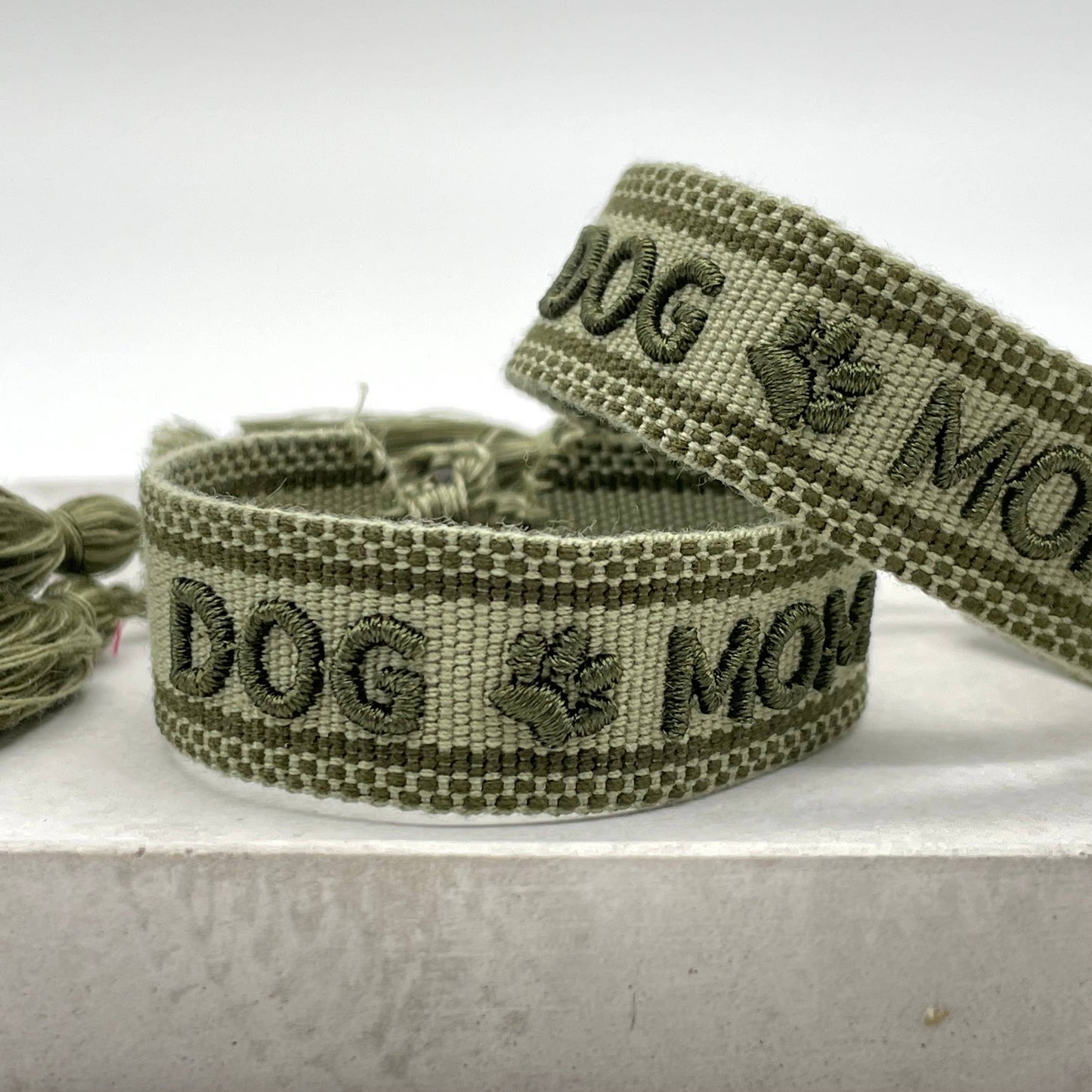 Grüner, gewebter Armreif mit der olivfarbener Aufschrift "DOG MOM". Der Armreif hat eine grobe Textur, und an den Enden befinden sich grüne Quasten.