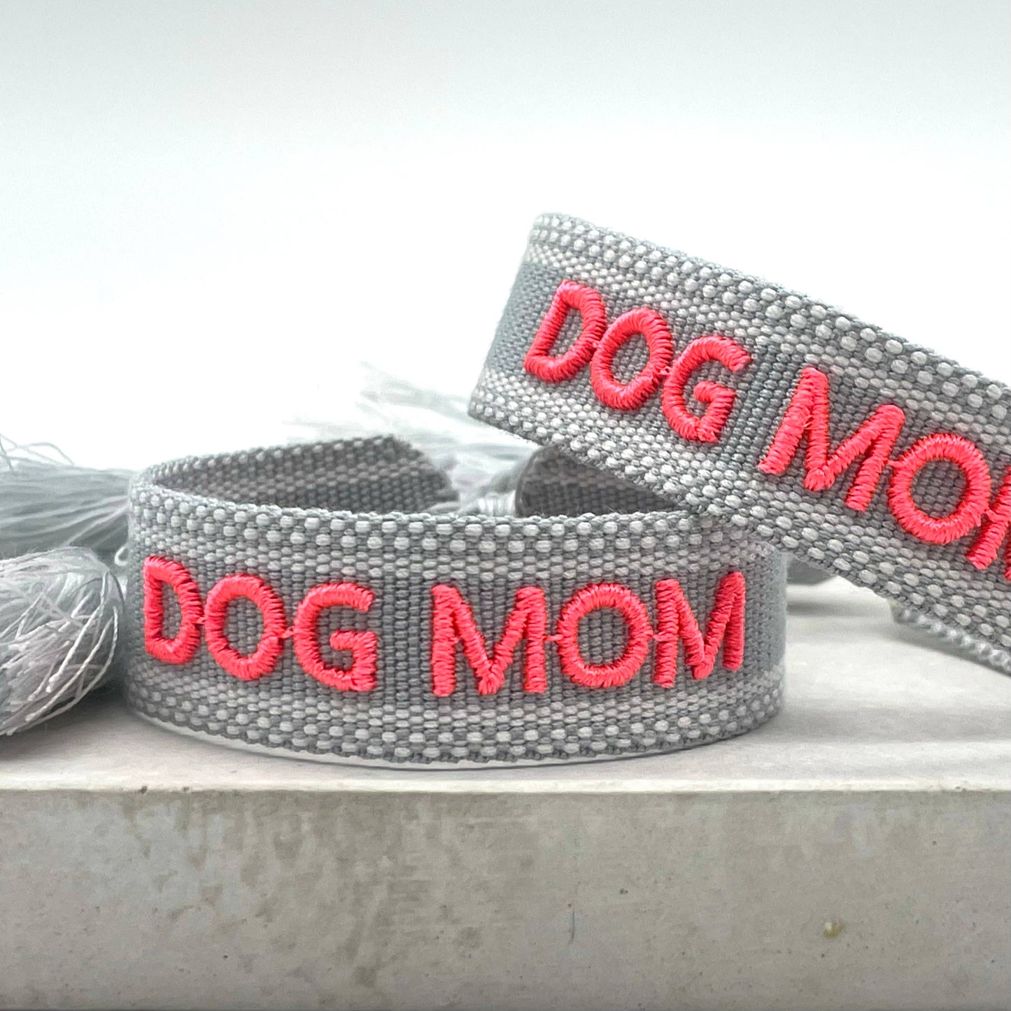 Grauer, gewebter Armreif mit der pinken Aufschrift "DOG MOM". Der Armreif hat eine grobe Textur, und an den Enden befinden sich graue Quasten.