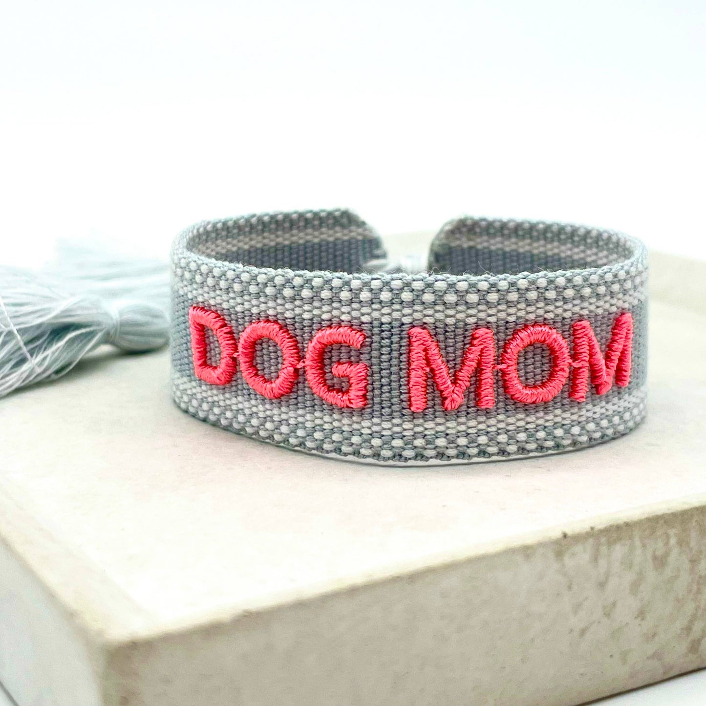 Graues, gewebtes Armband mit der pinkfarbenen Aufschrift "DOG MOM". Das Armband hat eine grobe, texturierte Oberfläche und an der Seite sind graue Quasten befestigt.