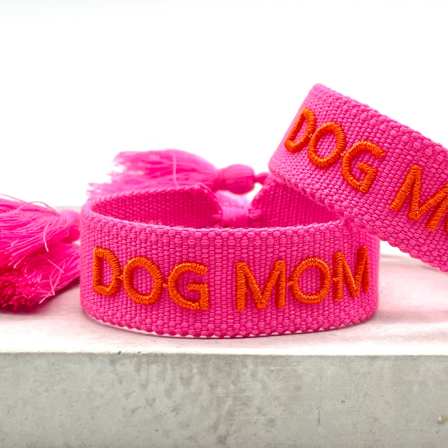 Pinker, gewebter Armreif mit der orangefarbenen Aufschrift "DOG MOM". Der Armreif hat eine grobe Textur, und an den Enden befinden sich pinke Quasten.