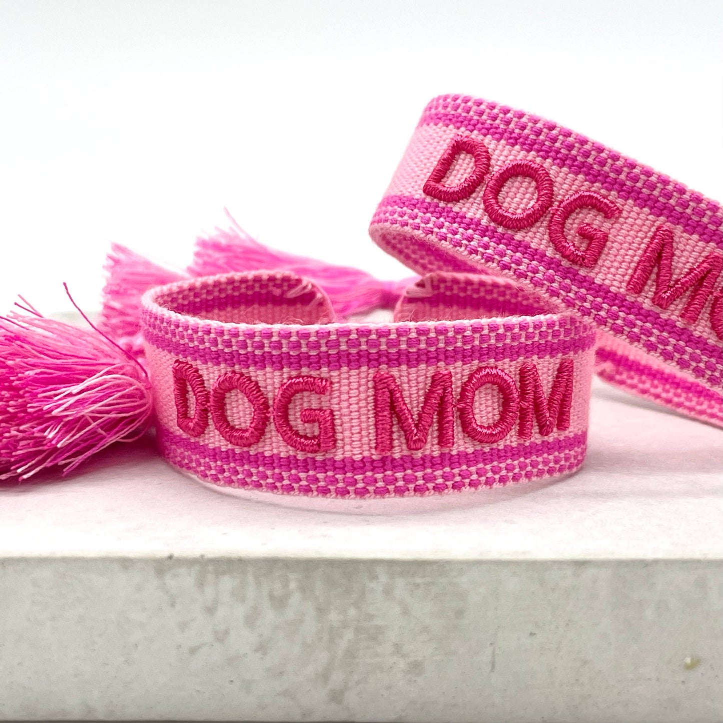 Rosa, gewebter Armreif mit der pinken Aufschrift "DOG MOM". Der Armreif hat eine grobe Textur, und an den Enden befinden sich rosa Quasten.