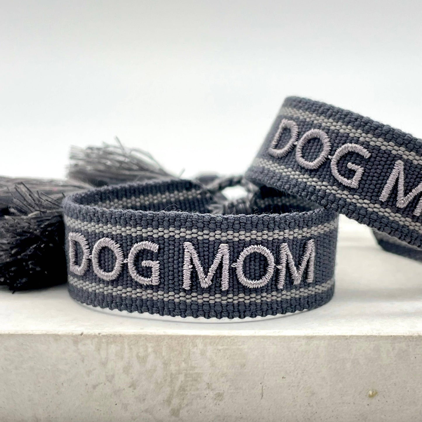 Schwarzer, gewebter Armreif mit der grauer Aufschrift "DOG MOM". Der Armreif hat eine grobe Textur, und an den Enden befinden sich schwarze Quasten.