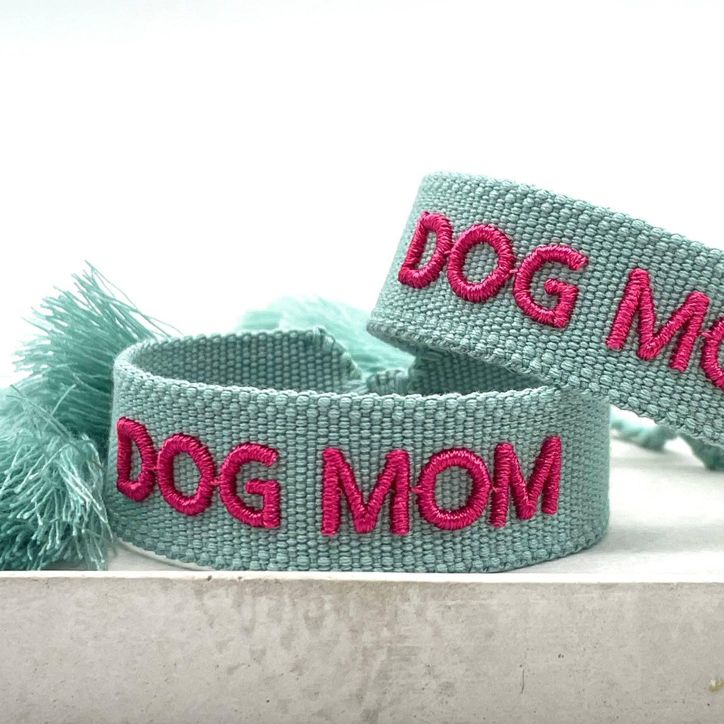 Türkiser, gewebter Armreif mit der pinken Aufschrift "DOG MOM". Der Armreif hat eine grobe Textur, und an den Enden befinden sich türkise Quasten.