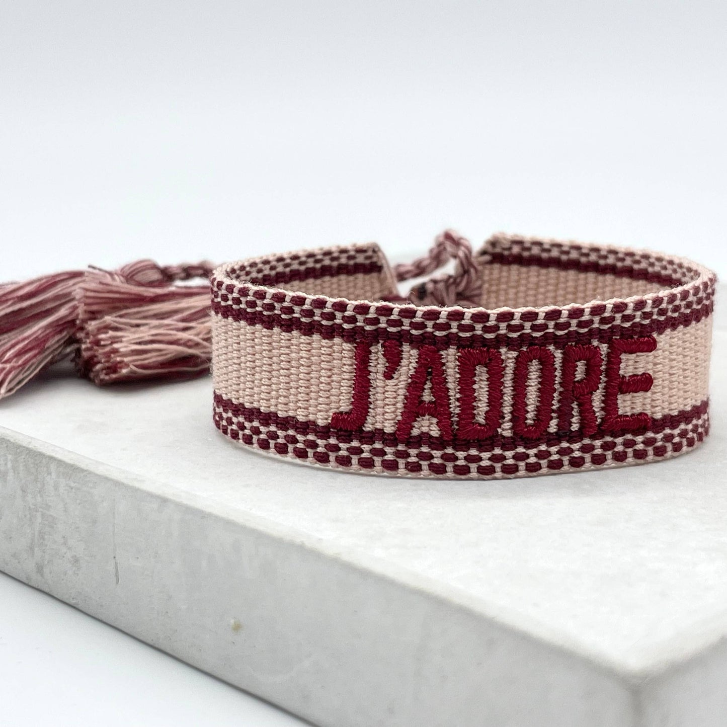 Gewebtes Armband in hellem rosa mit dunkelroter Aufschrift J'adore und farblich passenden Quasten. Das Armband liegt auf einem weißen Stein.