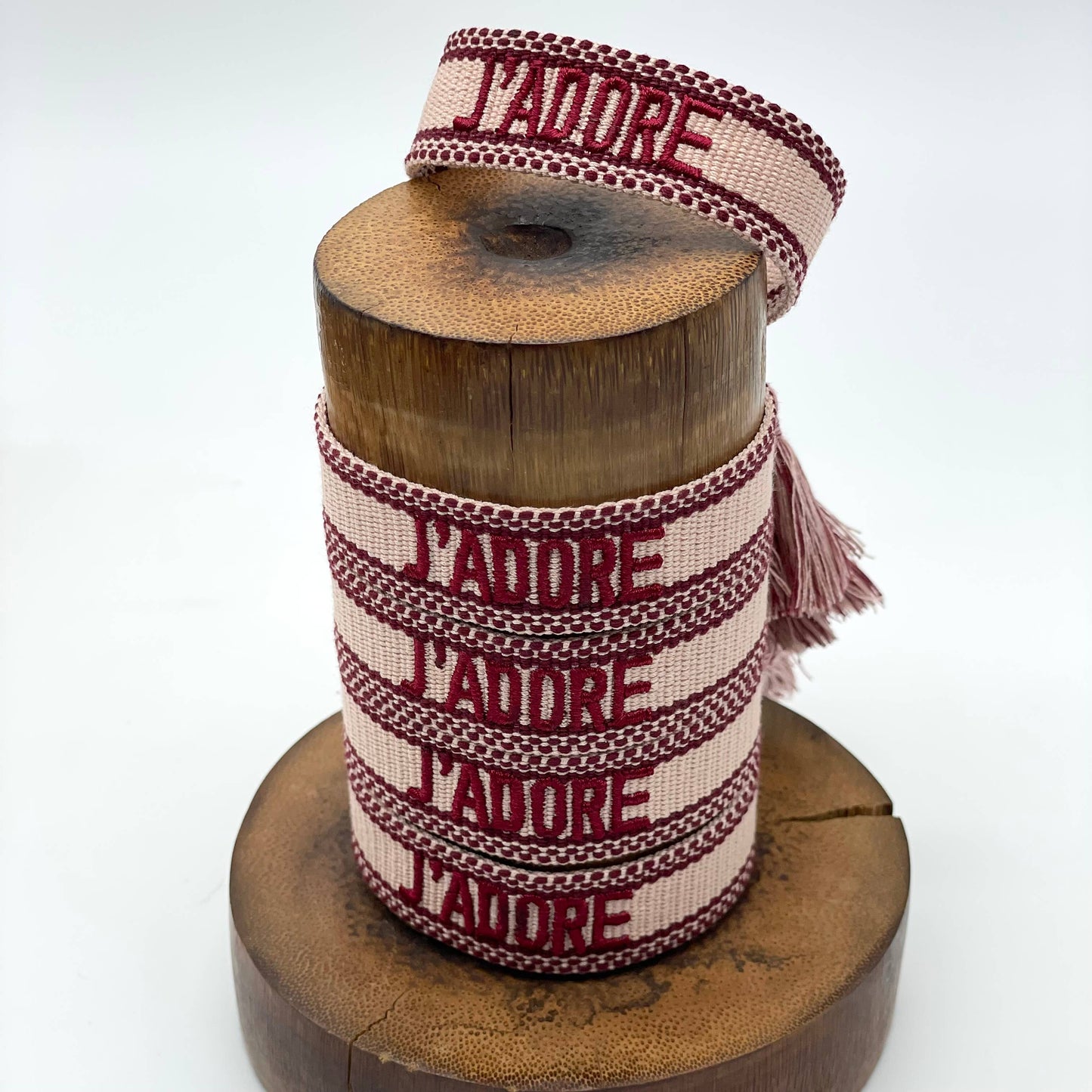 Mehrere Amrbänder in hellem rosa mit dunkelroter Aufschrift J'adore. alle gestapelt auf einem Holzständer.