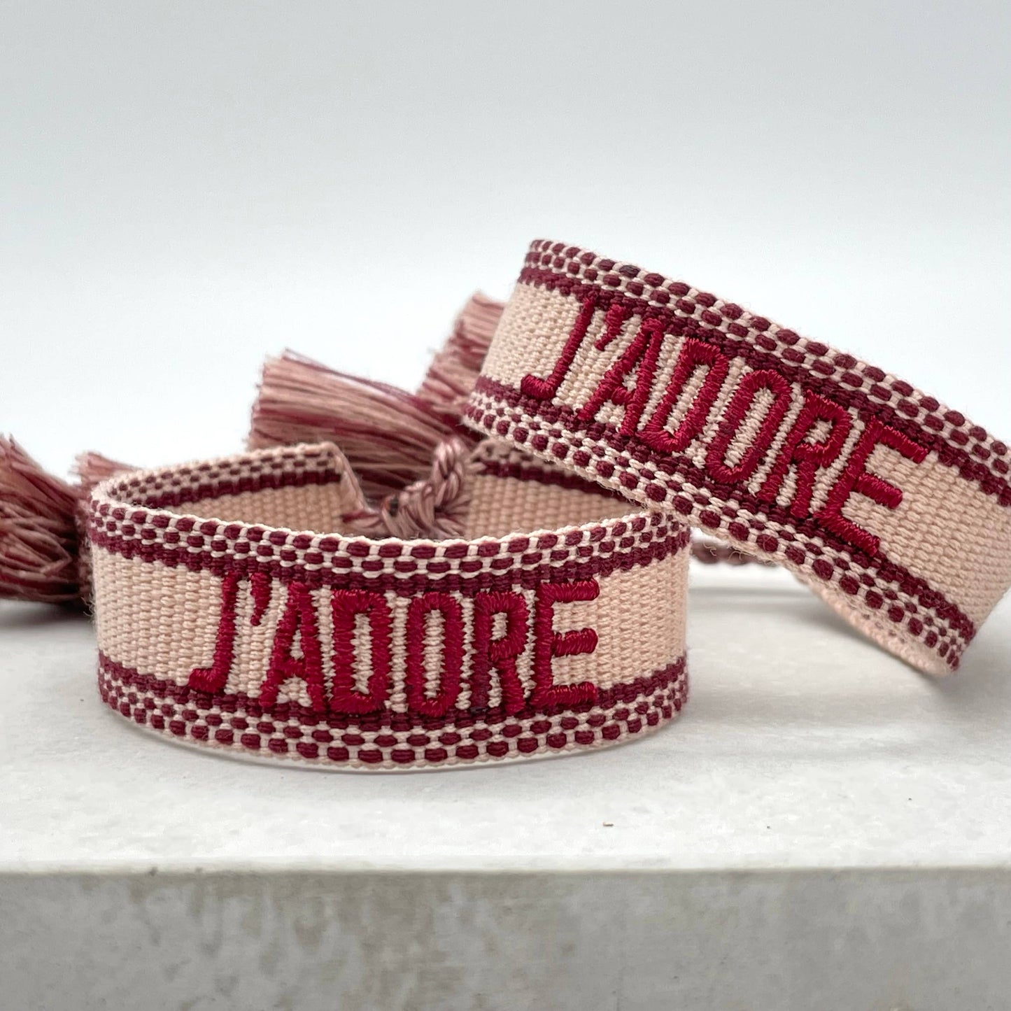 Beiges, gewebtes Armband mit der dunkelroten Aufschrift "J'ADORE". Das Armband hat eine strukturierte Oberfläche mit passenden Quasten an den Enden. Es handelt sich um ein elegantes Accessoire mit einem französischen Schriftzug, was "Ich liebe" bedeutet.
