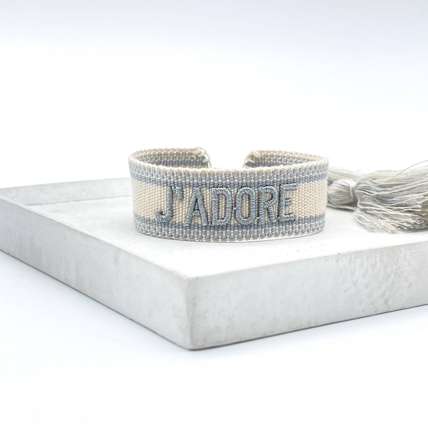 sandfarbenes, gewebtes Armband mit der silbernen Aufschrift "J'ADORE". Das Armband hat eine strukturierte Oberfläche mit passenden Quasten an den Enden. Es handelt sich um ein elegantes Accessoire mit einem französischen Schriftzug, was "Ich liebe" bedeutet.