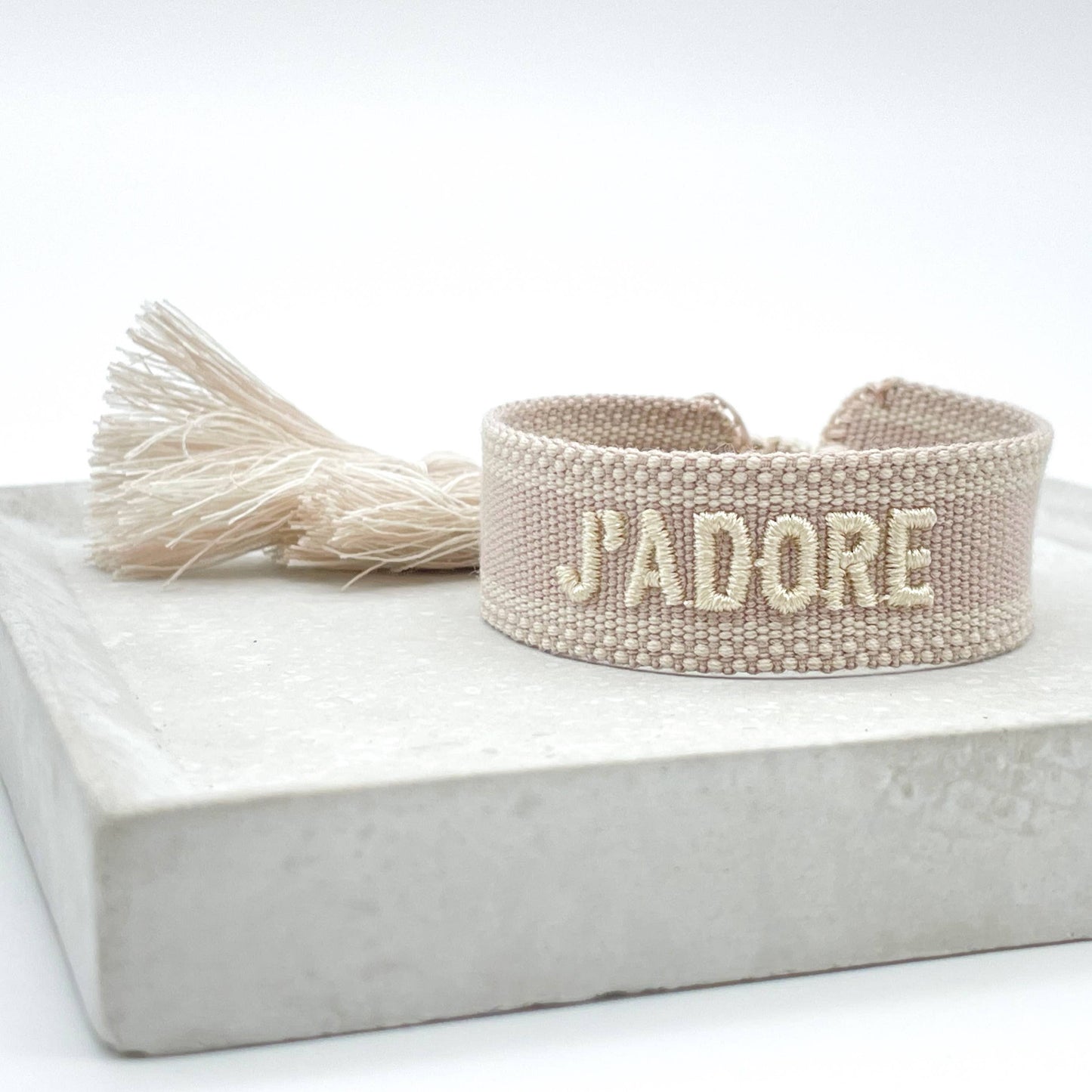 Beiges, gewebtes Armband mit der helleren Aufschrift "J'ADORE". Das Armband hat eine strukturierte Oberfläche mit passenden Quasten an den Enden. Es handelt sich um ein elegantes Accessoire mit einem französischen Schriftzug, was "Ich liebe" bedeutet.