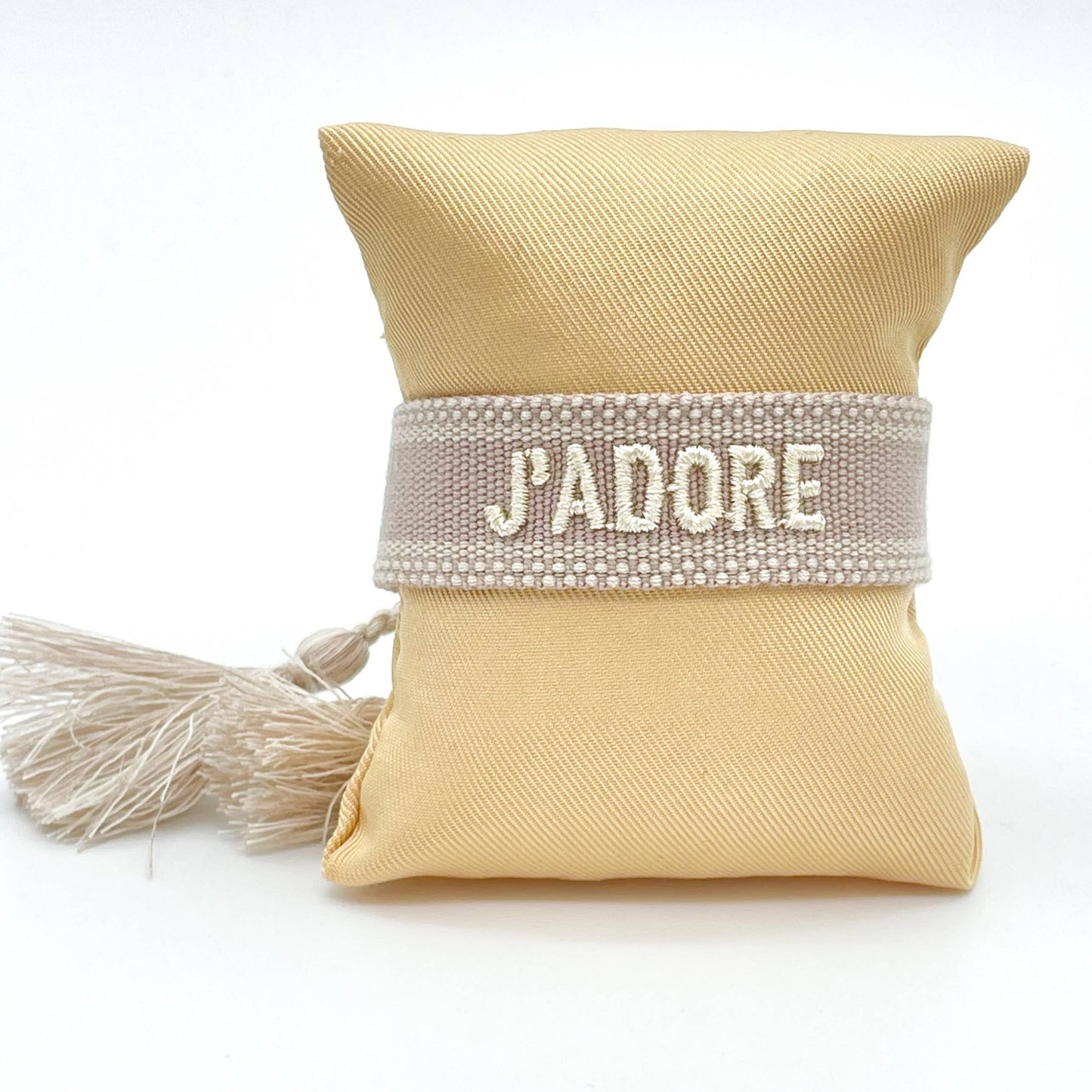 Gewebtes Armband in beide mit heller Aufschrift J'adore und beigen Quasten. Das Armband befindet sich auf einem Kissen.