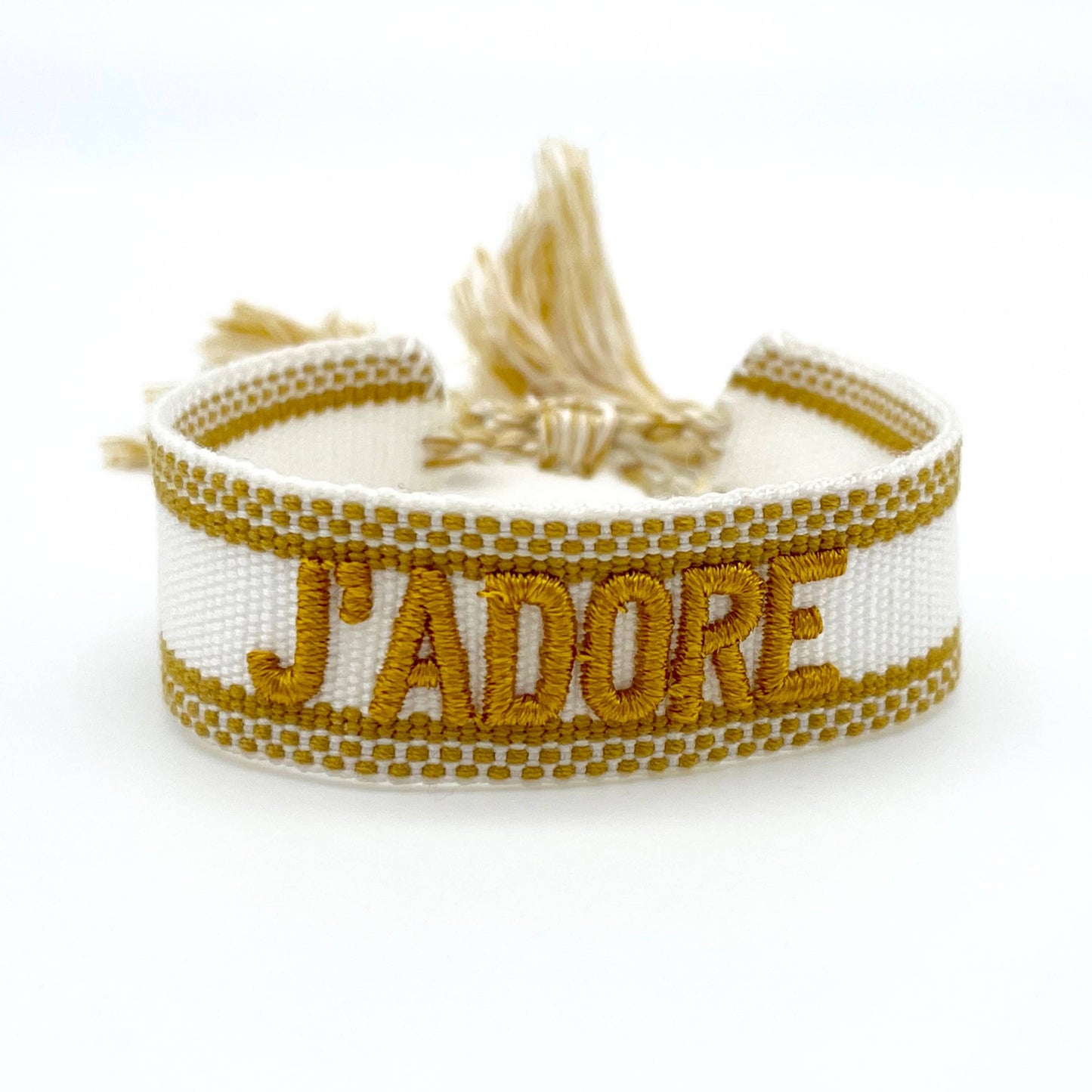 Weißes, gewebtes Armband mit der goldenen Aufschrift "J'ADORE". Das Armband hat eine strukturierte Oberfläche mit passenden Quasten an den Enden. Es handelt sich um ein elegantes Accessoire mit einem französischen Schriftzug, was "Ich liebe" bedeutet.