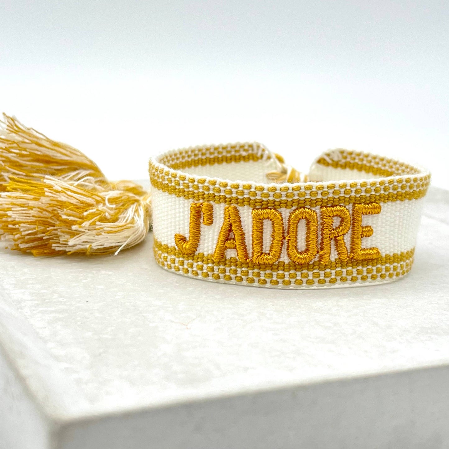Gewebtes Armband in weiß mit goldener Aufschrift J'adore und farblich passenden Quasten. Das Armband liegt auf einem weißen Stein.