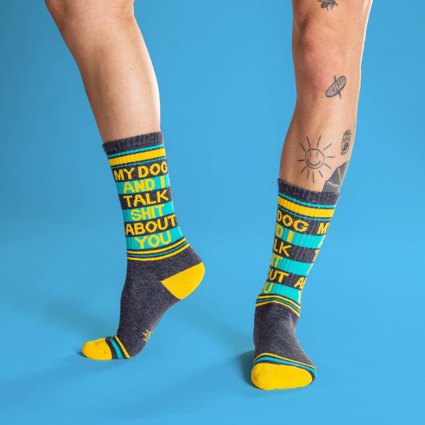 Ein Paar Socken in den Farben Dunkelgrau, gelb und Türkis. Die Aufschrift sagt My dog an I talk shit about you. Sie werden von einem Mann getragen, die Beine sind ab Knie abwärts zu sehen.
