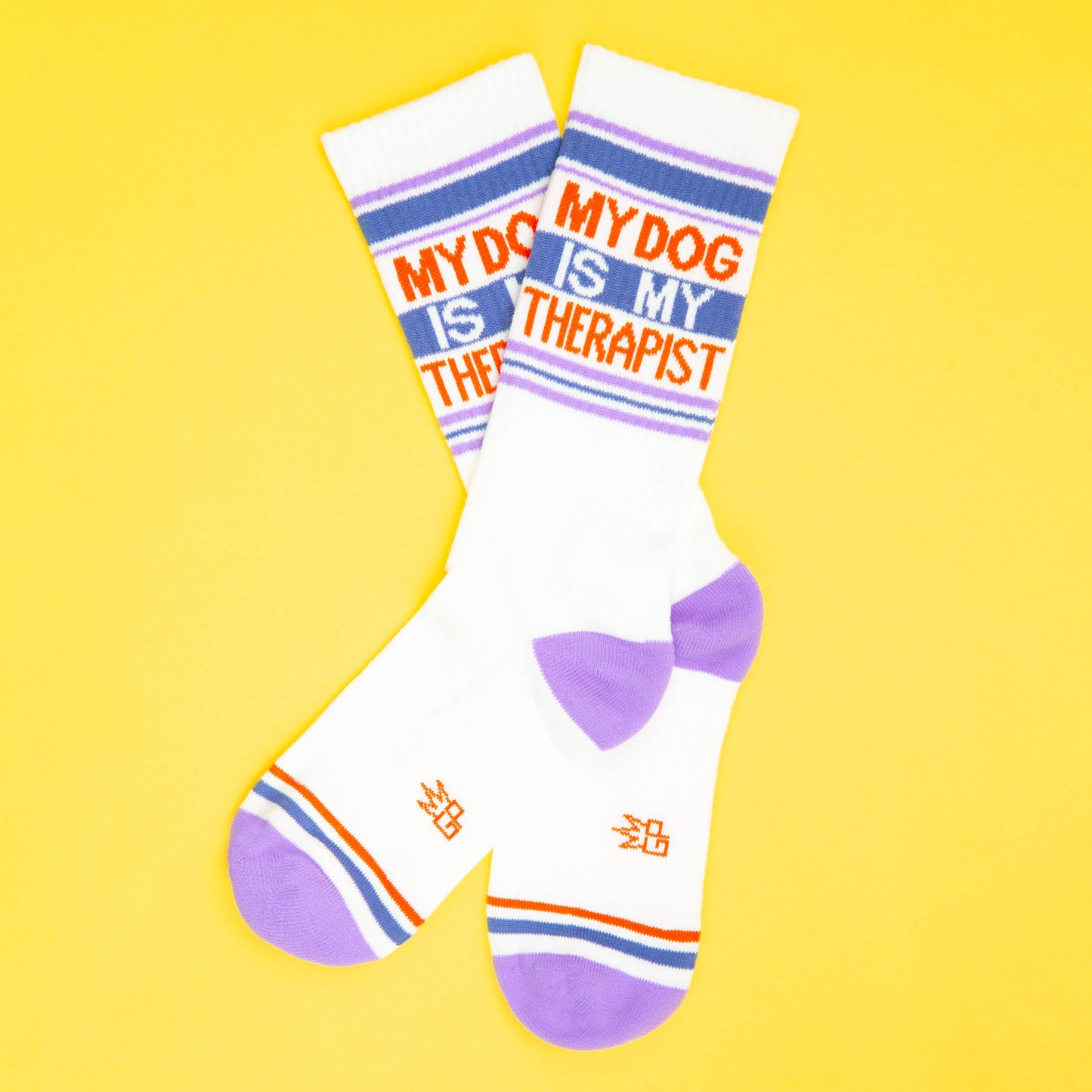 Ein Paar Socken inden Farben weiß mit blau, rot und Flieder. Die Aufschrift sagt my dog is my therapist. Beide Socken liegen übereinander auf gelebem Untergrund.