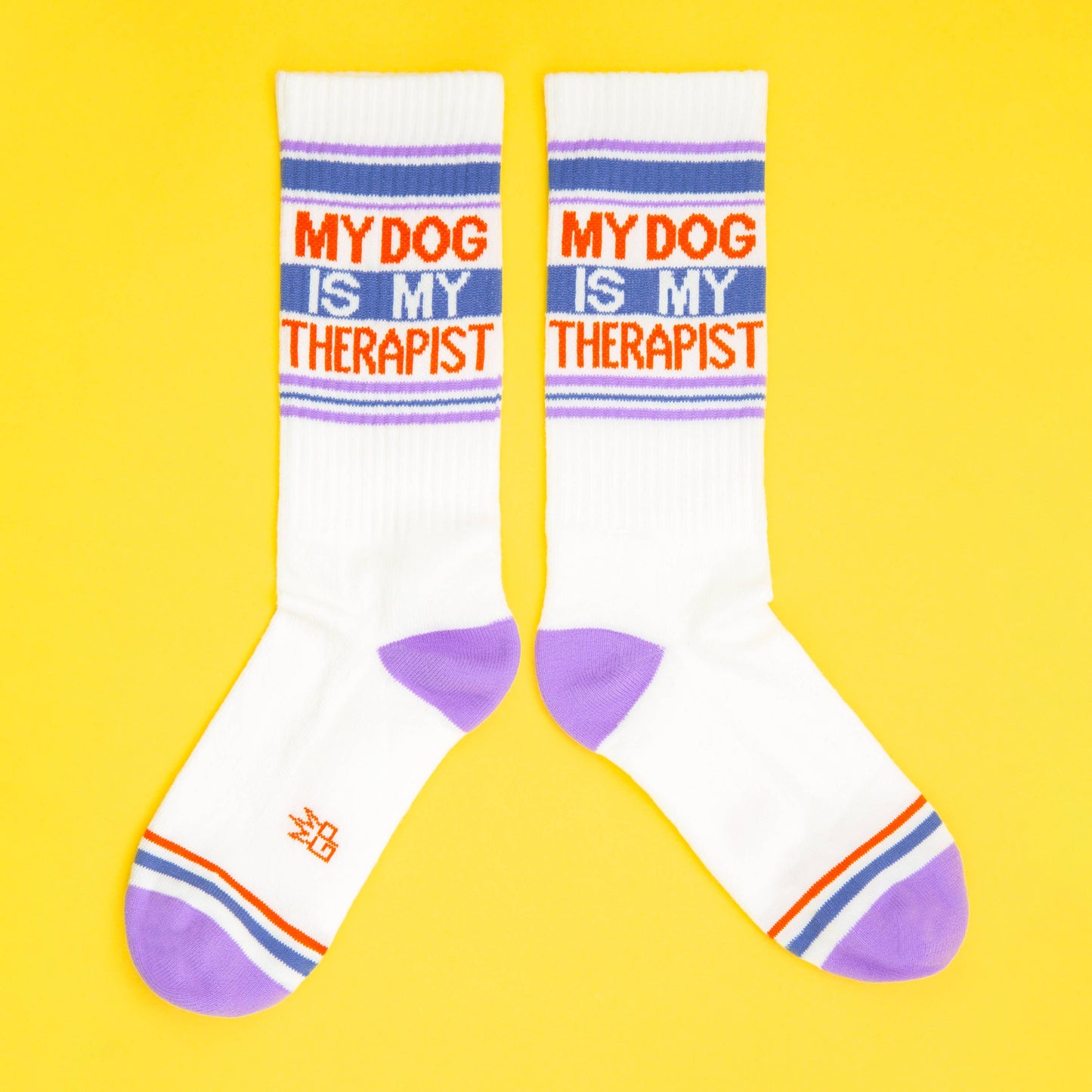 Ein Paar Socken inden Farben weiß mit blau, rot und Flieder. Die Aufschrift sagt my dog is my therapist. Die Socken sind von Gumball Poodle. Sie leige auf gelbem Untergrund und zeigen in unterschiedliche Richtungen.