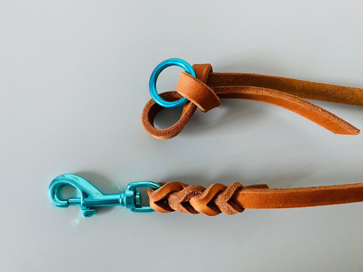 Beide Enden der Lederleine, einmal Knoten mit Ring und einmal Karabiner mit Flechtung. Beide Beschläge in Türkis, das Leder in Cognac.