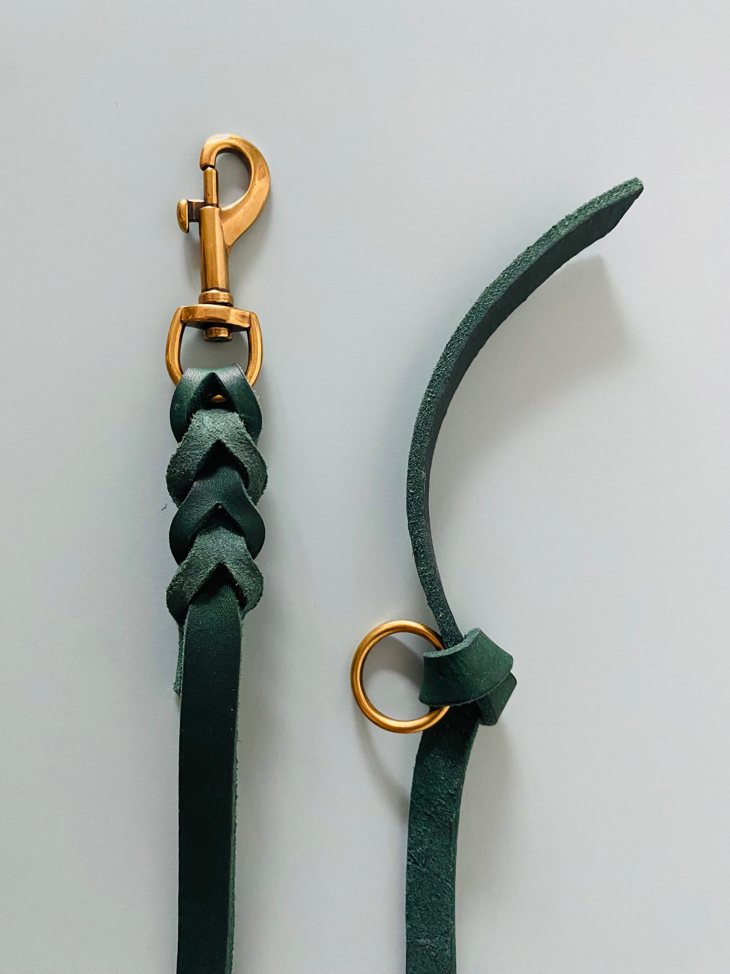 Zwei Enden einer Hundeleine aus Leder in dunkelgrün. Auf der einen Seite ien Karabiner in gold , der in das Leder eingeflochten ist. Auf der anderen Seite ein Knoten mit Ring in Gold.