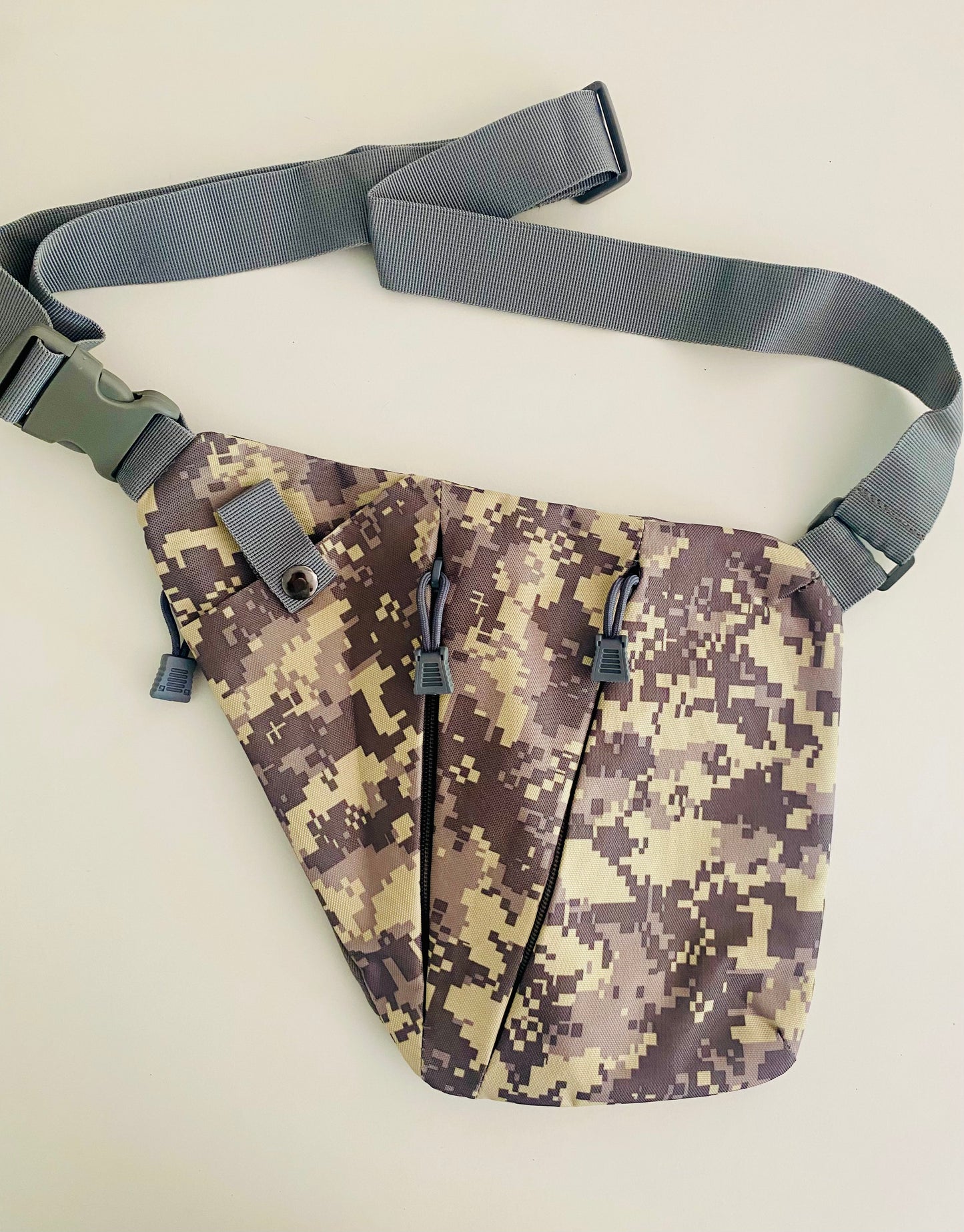 Tasche aus Canvas Stoff in Camouflage. Die Tasche hat zwei Reißverschlüsse und ein Fach, das mit einem Koopf verschlossen ist. Die Tasche verfügt über einen Träger, der längenverstellbar ist.