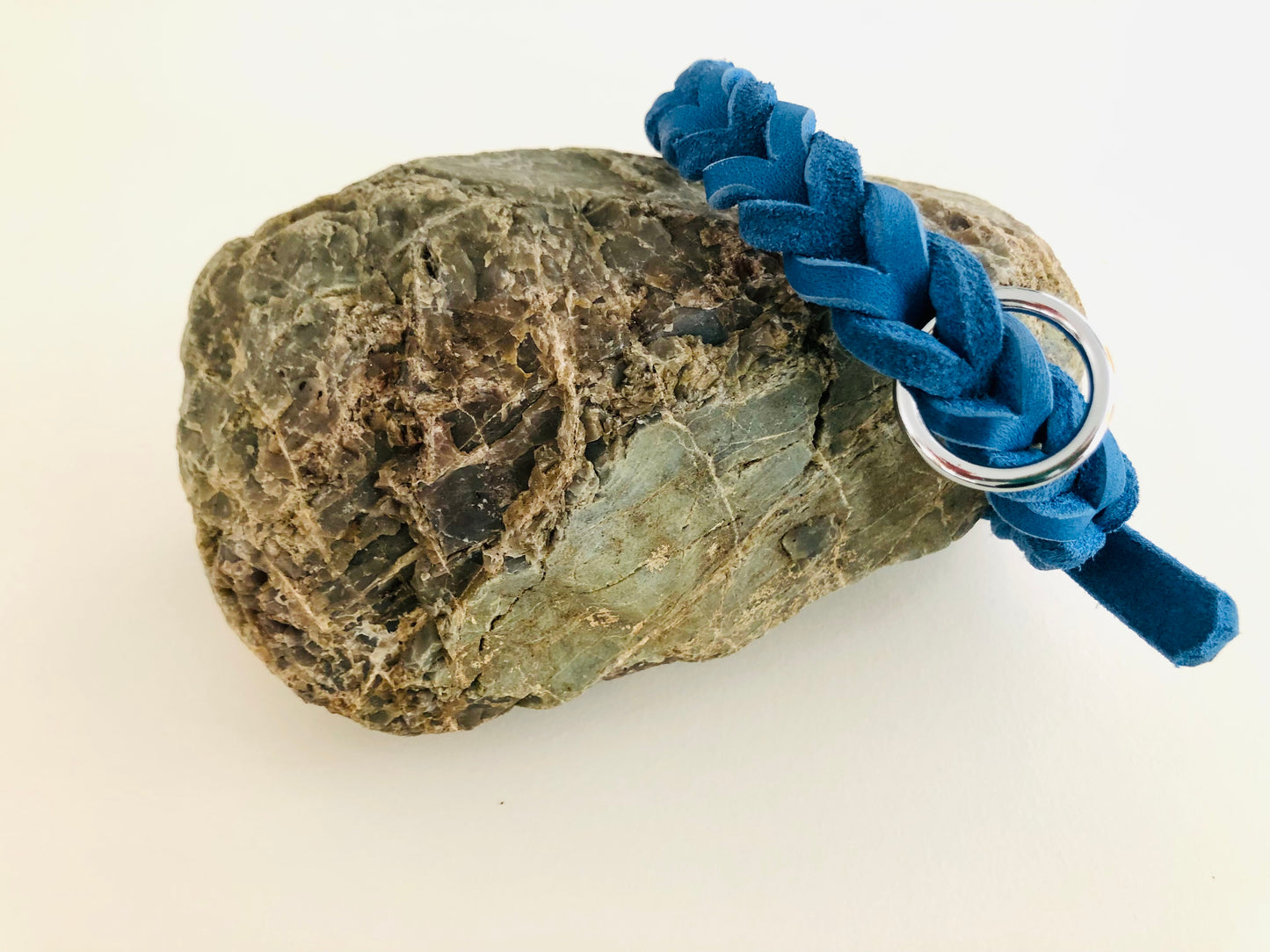 Ein blaues, geflochtenes Fettlederhalsband mit silberner Schnalle und Metallring, auf einem Stein liegend.