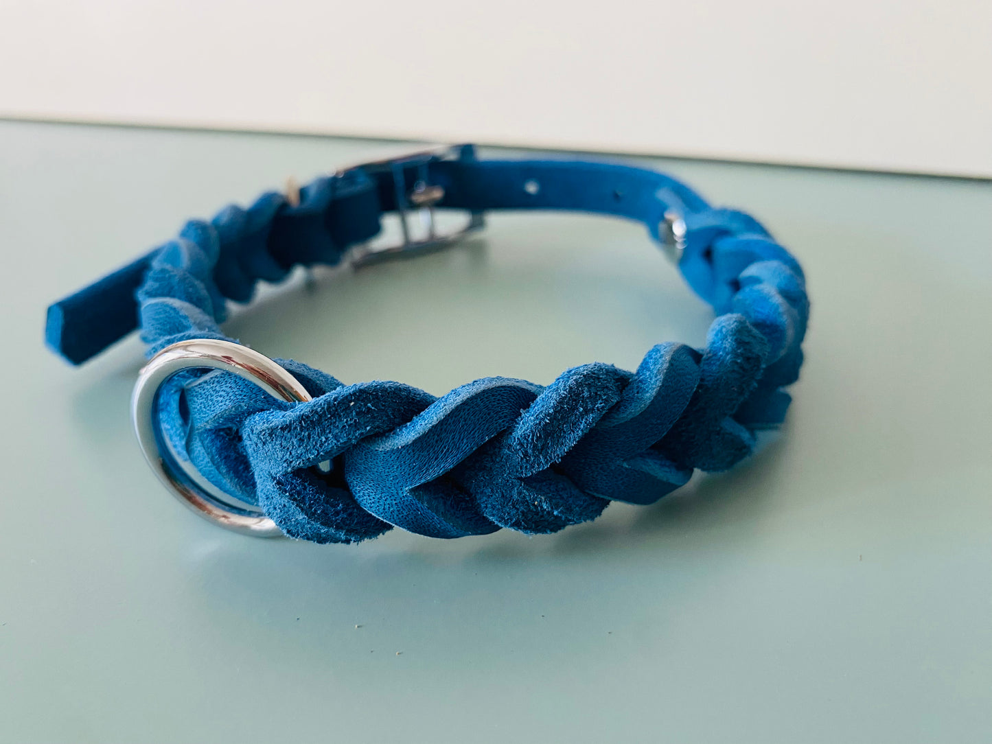 Ein blaues, geflochtenes Fettlederhalsband mit silberner Schnalle und Metallring, seitlich fotografiert auf hellem Untergrund.