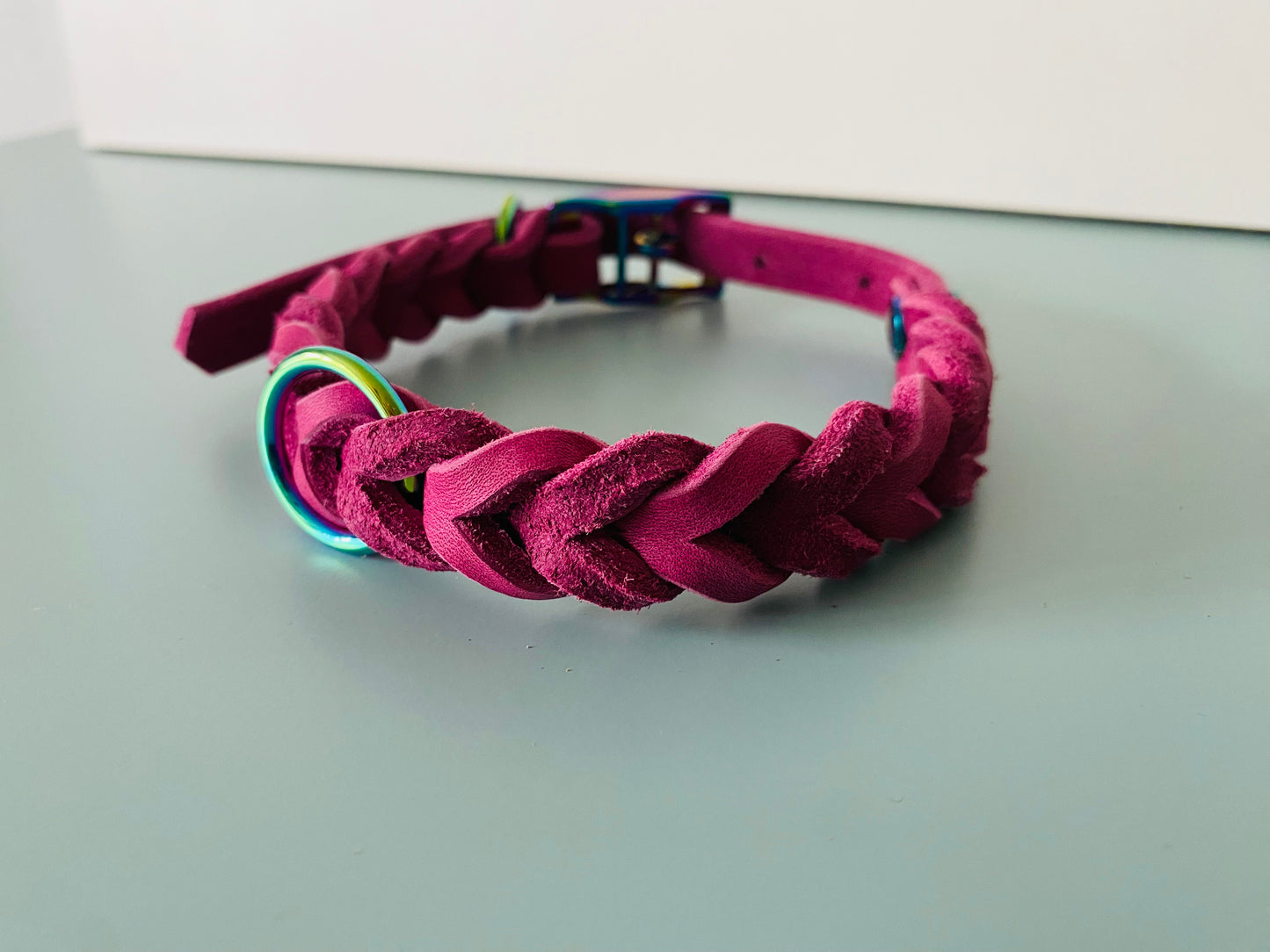 Ein magentafarbenes, geflochtenes Wildlederarmband mit einem regenbogenfarbenen Metallring und einer passenden Schnalle, vor einem magentafarbenen Hintergrund.“