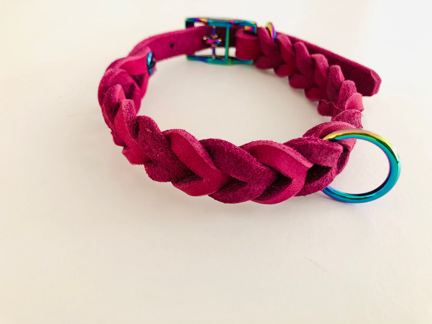 Ein magentafarbenes, geflochtenes Fettlederhalsband mit einem regenbogenfarbenen Metallring und einer passenden Schnalle, auf hellem Untergrund liegend.