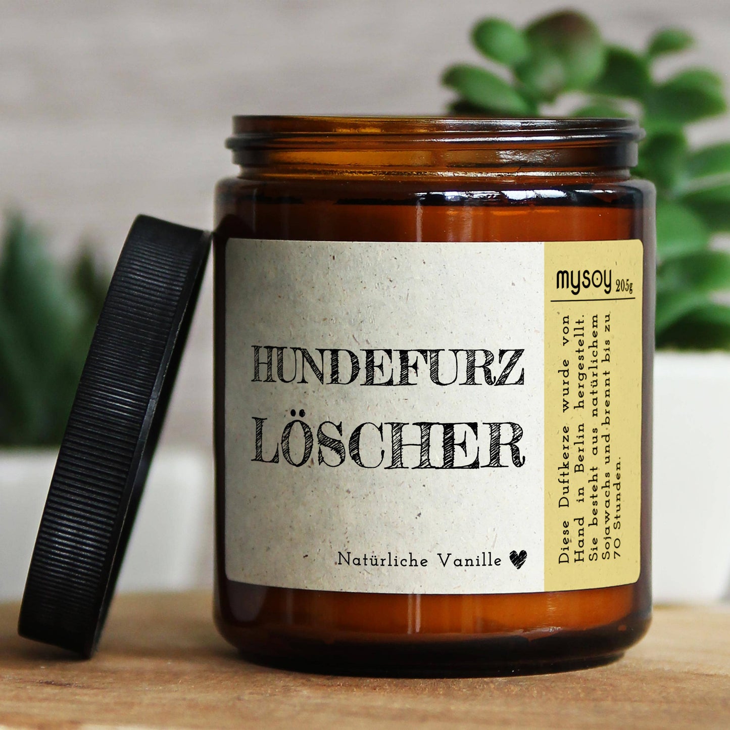 Dunkelbraunes Glas mit schwarzem Deckel, der danbene liegt. Die Aufschrift sagt Hundefurzlöscher, es ist ein eDuftkerze mit dem Aroma Natürliche Vanille.