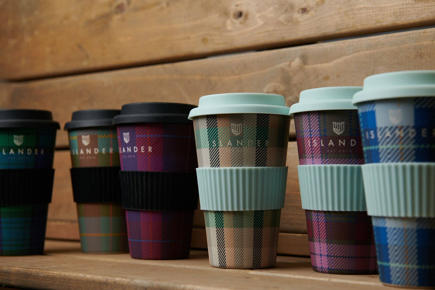 Mehrere Thermobecher mit Tartanmuster in verschiedenen Farben . Die Becher haben einen Deckel und eine Banderole aus Kunststoff.